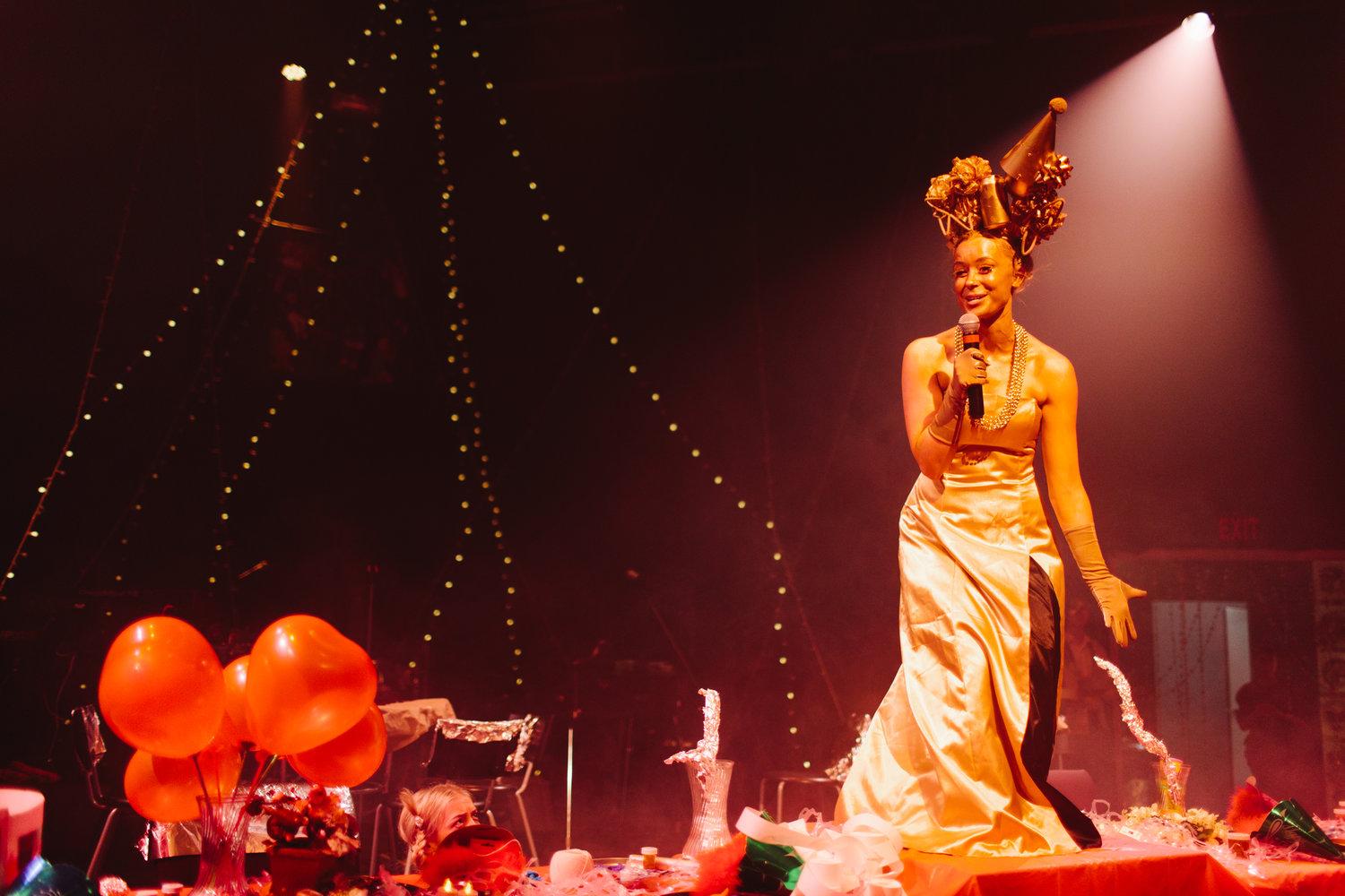 A performer on stage wearing a gown with extravagant head piece speaking to the audience while having starry lighting strings and balloons in the background