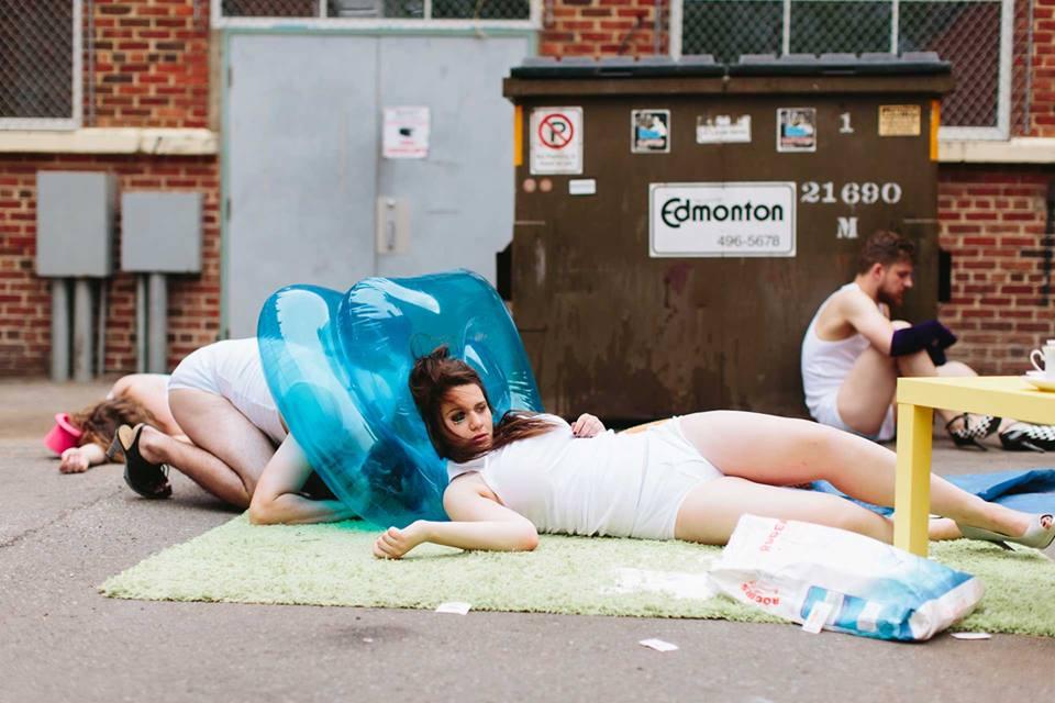 Setting in the outside of the back of the building and a commercial waste bin in the daytime. There are 4 people laying on the ground in their under clothes. In the center two people leaning against an inflatable blue plastic floaty while a man sitting and holding his knees close to his body. They are all wearing heels