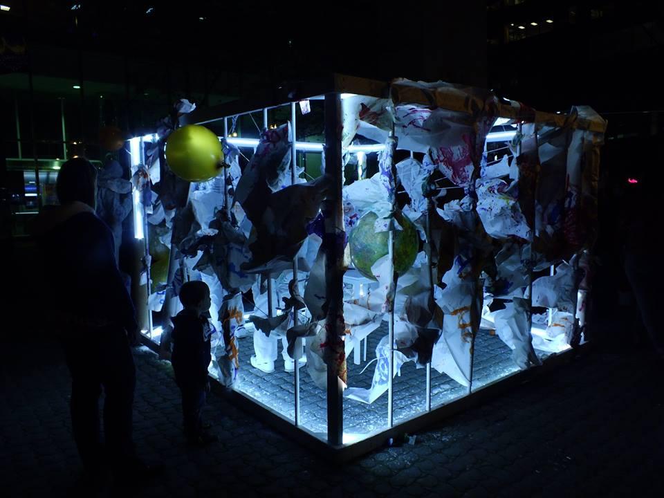 Public installation of a cage structure with loose paper and balloon around the exterior. The led strips on the inside illuminated installation at night