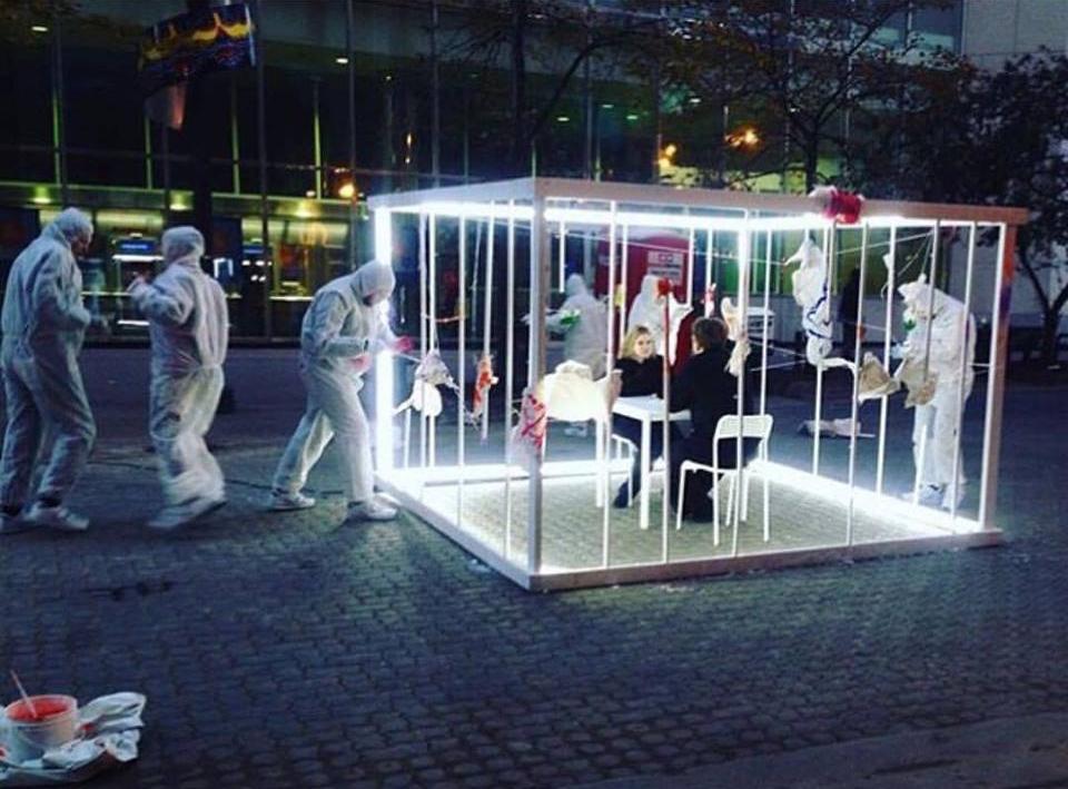 A white cage like installation with a couple wearing all black sitting in front of each other while six people wearing body protection suit seemingly busy in the exterior of the installation