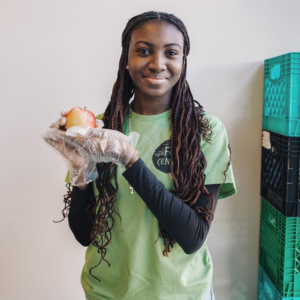 An SCSU food centre volunteer in a light green volunteer shirt holding up an apple in gloved hands
