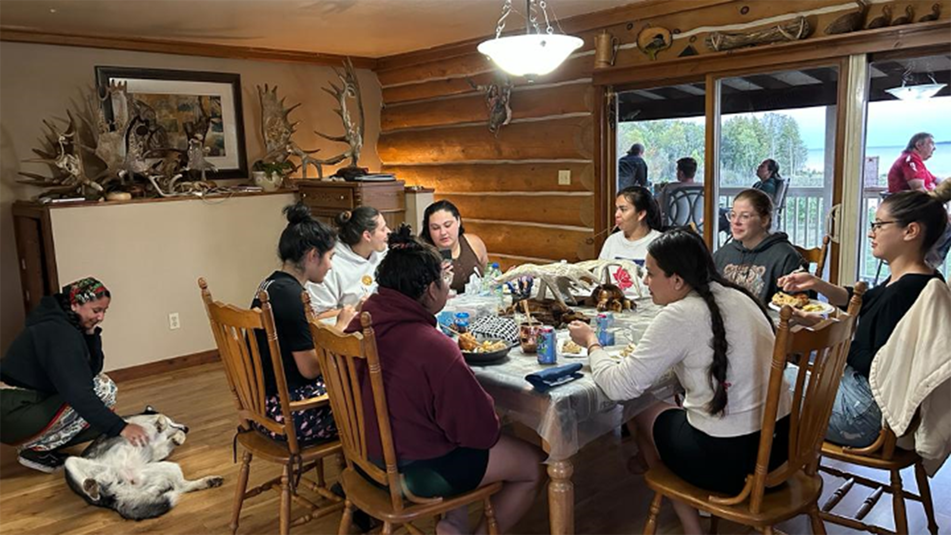 8 people sitting at a dining table enjoying a dinner feast, with a husky lying on their back getting their belly rubbed by a participant