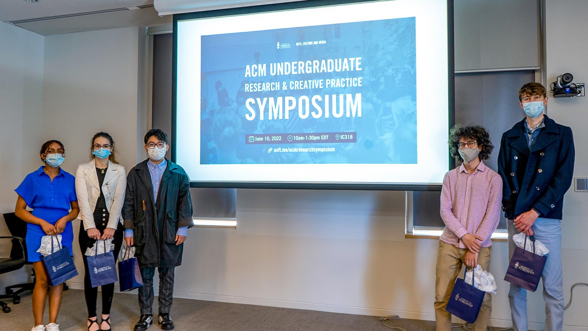 Group photo of ACM student presenters standing in front of the projector with the banner of the Symposium