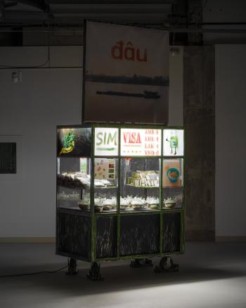 Art installation of a food cart with a sign that says SIM, VISA, RMB, KHR, LAK, VND. Inside the cart has metal bowls containing sim cards and some dehydrated food.
