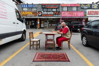 Reza Nik is sitting in a parking lot for a durational performance piece and has setup a table and another chair across from him