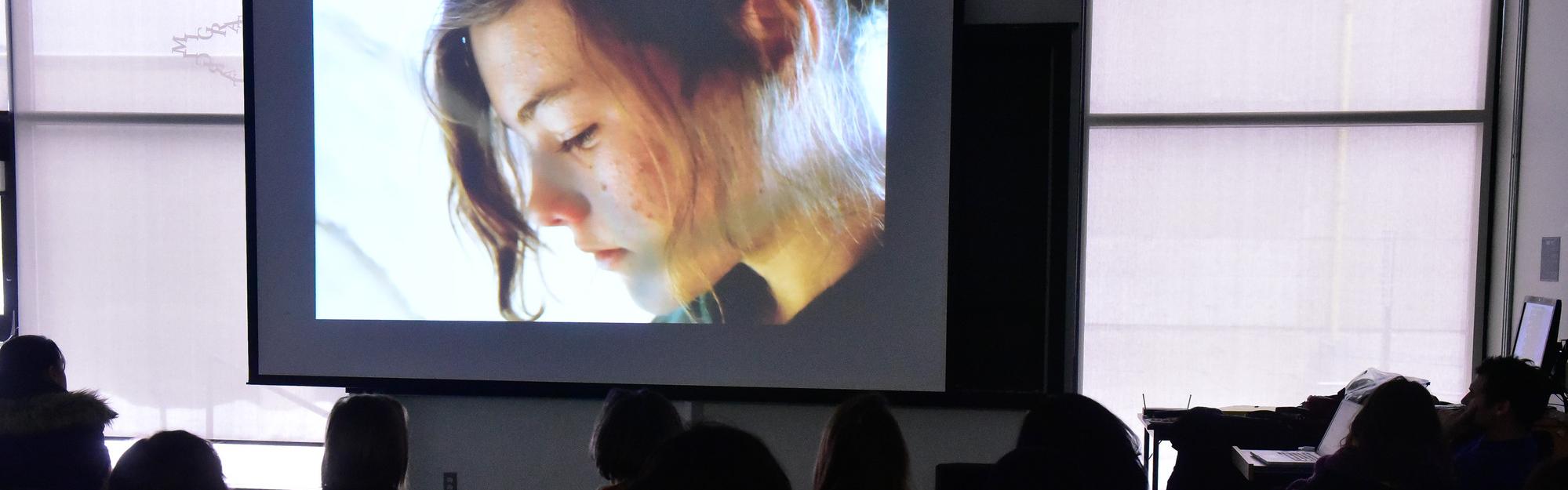 A still from a film, close shot of a young woman's face, with students watching the screen in the foreground