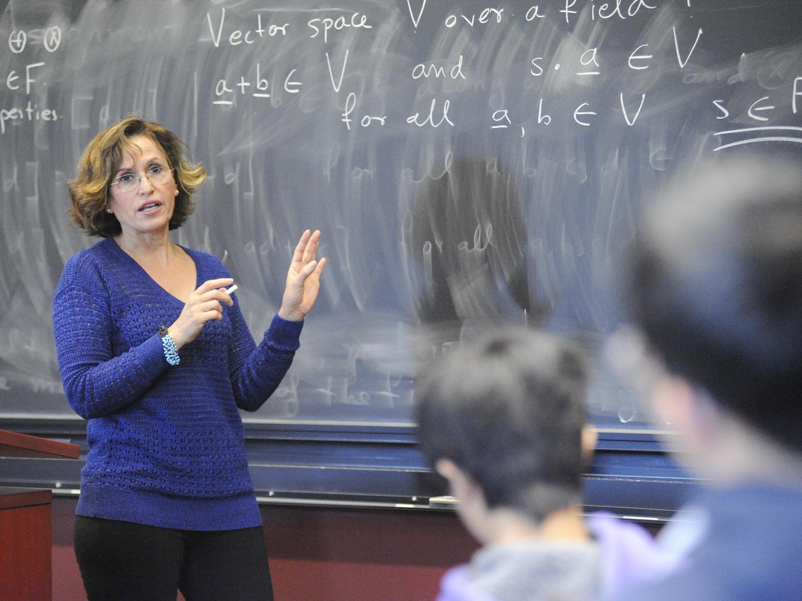 A professor in front of a blackboard with mathematical expressions behind, and students in the foreground