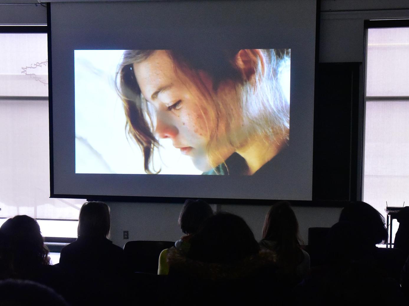 A still from a film, close shot of a young woman's face, with students watching the screen in the foreground