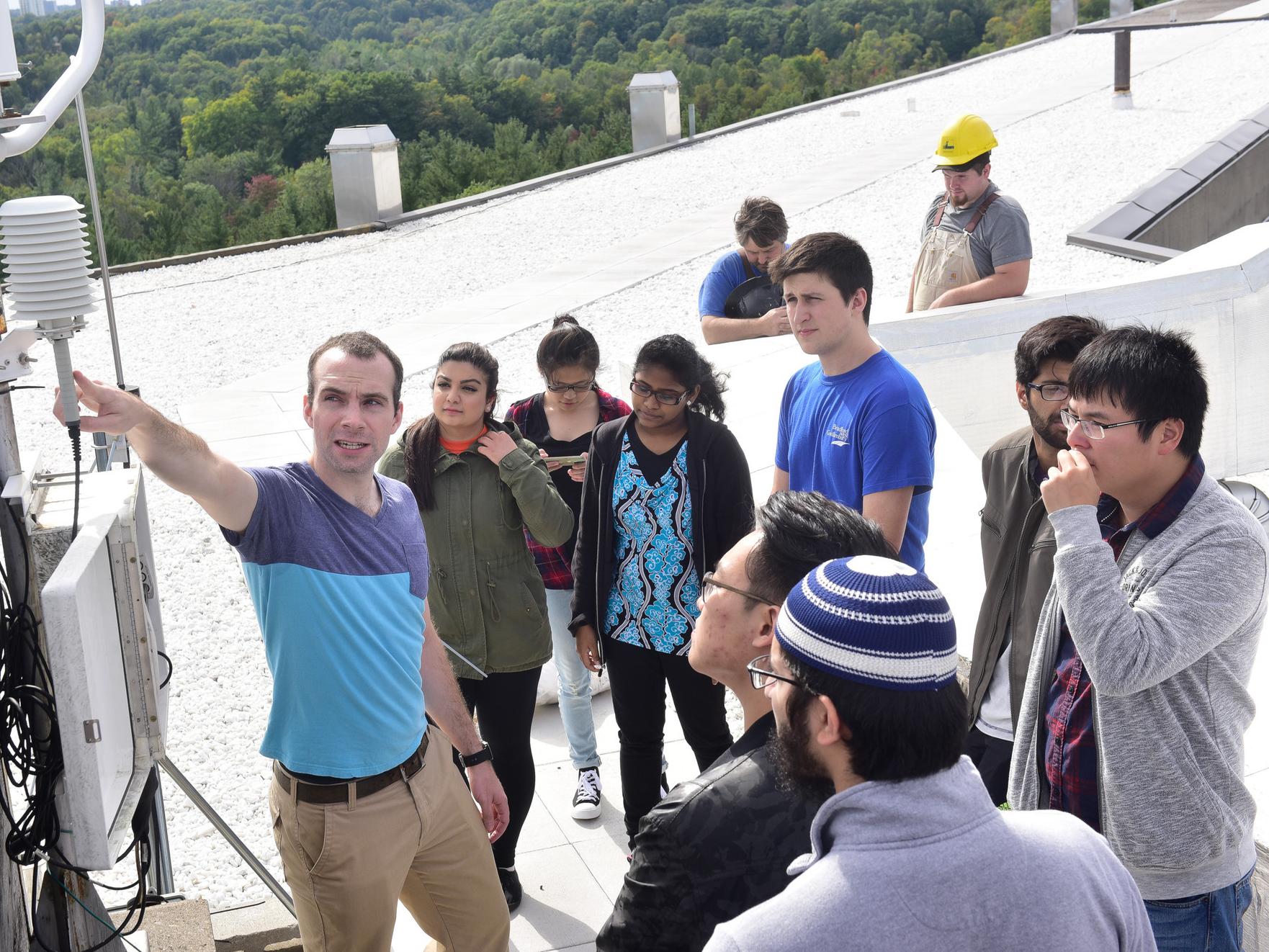 A group of students on a field trip to a weather station, on a roof examining a weather vane