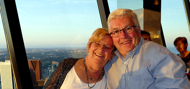 Stephanie and Bruce Geddes lean into one another, smiling, with the backdrop of the city behind them