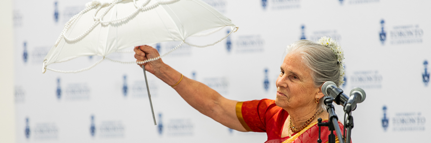 Professor Brenda Beck holding up a white pearled umbrella in front of a step and repeat with UTSC logo