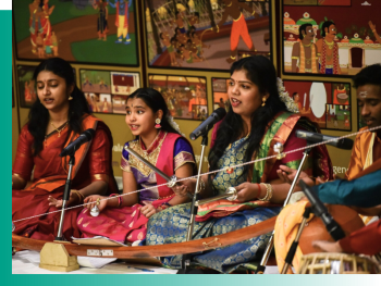 three singers in front of a mic in very colourful traditional saris, with a person playing the tabla in the right corner