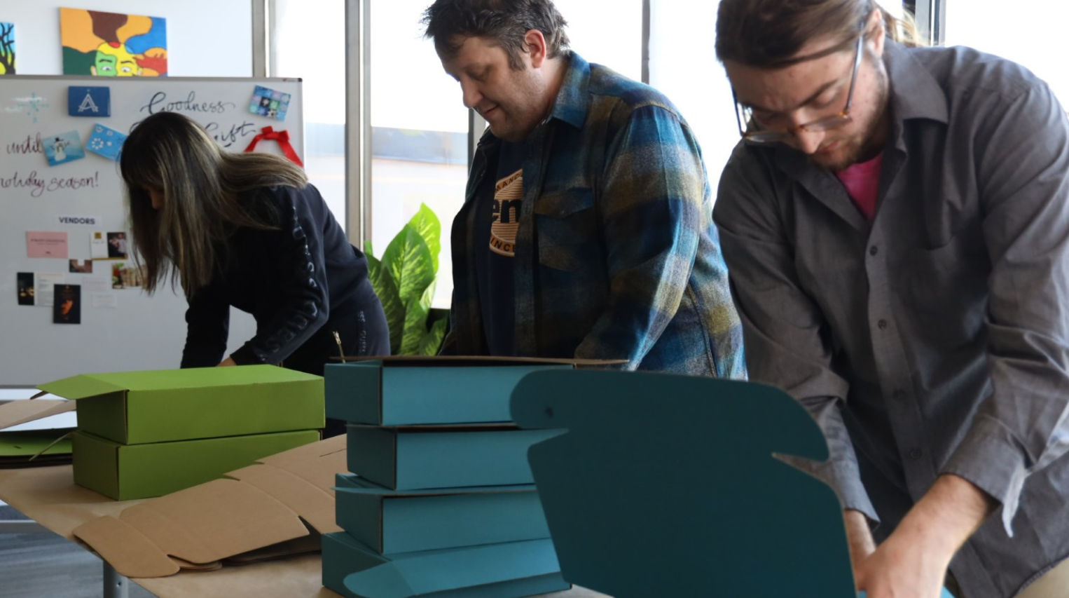 Three people are actively engaged in assembling gift boxes in a bright, indoor setting. Stacks of flat, teal and green box pieces are being crafted into three-dimensional shapes on a long table. A display board in the background features a cheerful notice about the holiday season, suggesting a festive, community-driven event.