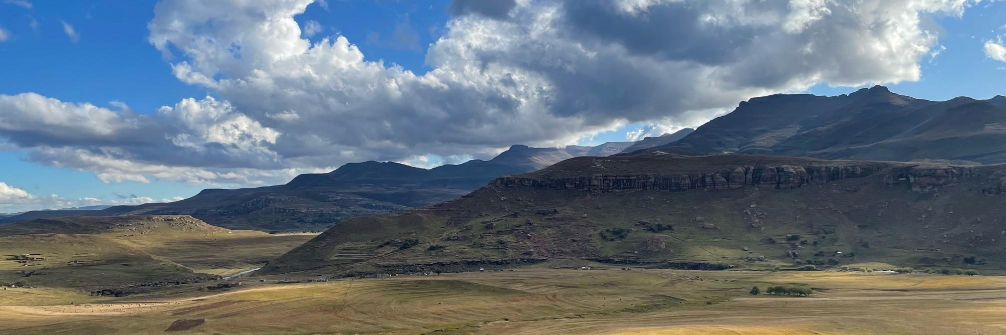 A vista of the Maloti-Drakensberg mountain area in Lesotho. A rolling plain gives way to tall, craggy mountains under blue skies with white puffy clouds.