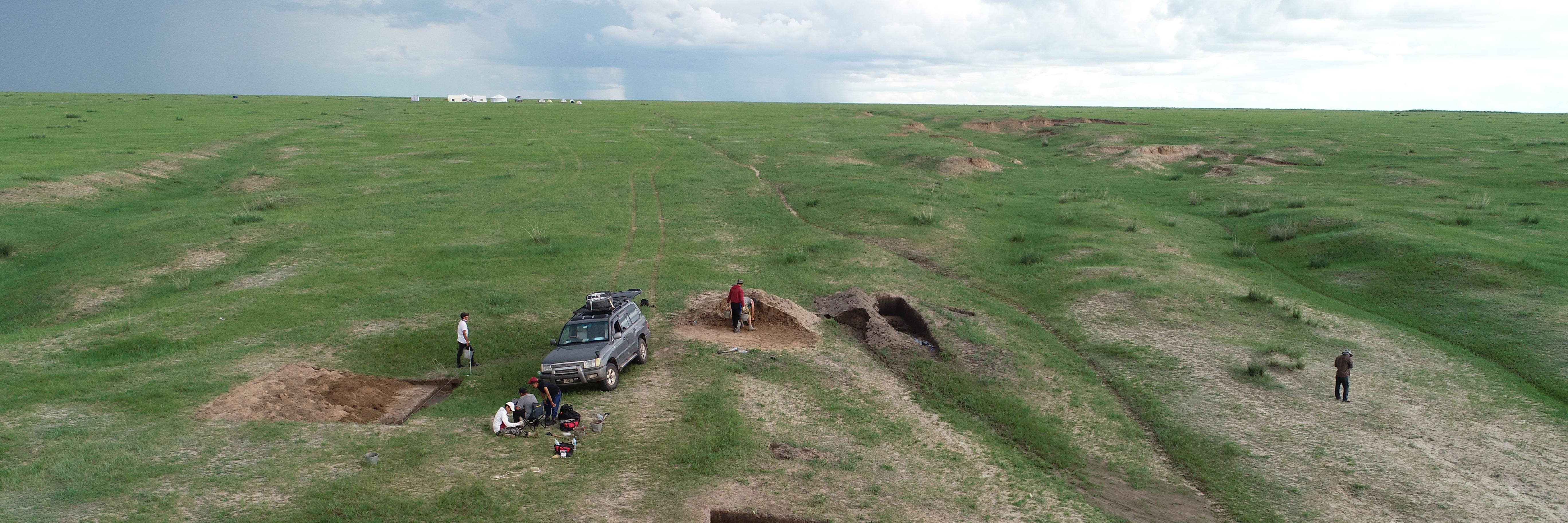 Archaeologists work on excavating a home in the foreground, with a wide expanse of steppe behind