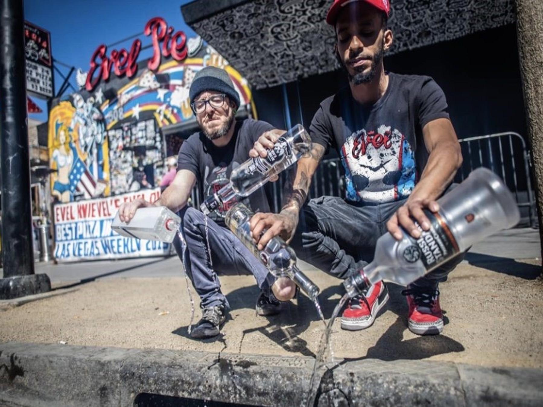 Two punk rockers pouring out bottles of vodka in the gutter, graffiti wall background
