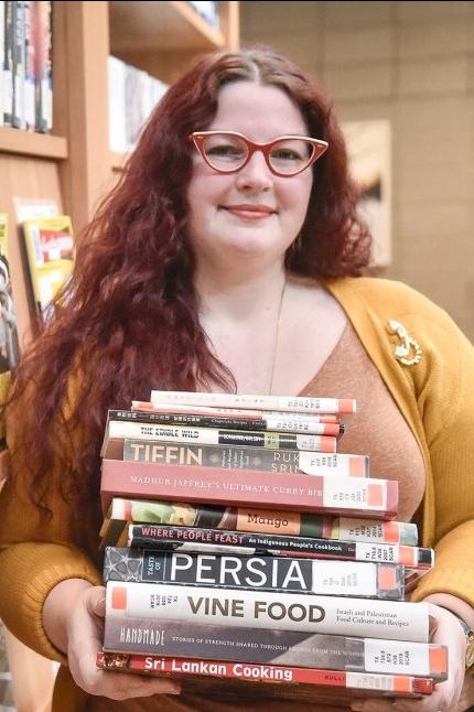 Whitney Kemble wearing red framed glasses and a yellow cardigan holding a stack of cookbooks and food studies-related books with both hands