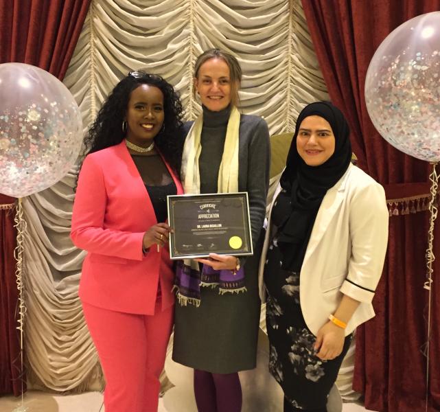 Dr. Bisaillon, a tall white woman, receives an award from two women of color, while standing on a stage wtih balloons and a festive backdrop