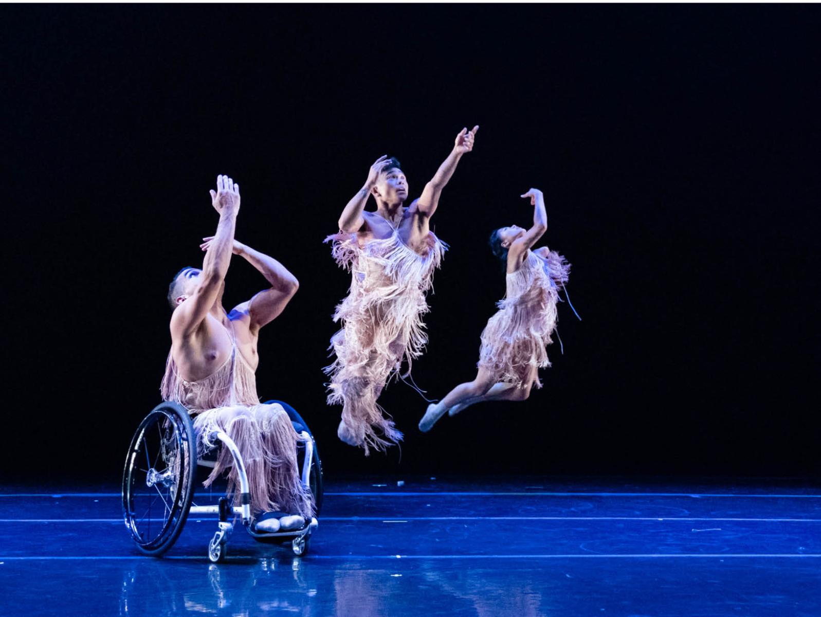 Three dancers in feathery costumes on stage, one is seated in wheel chair, others leap