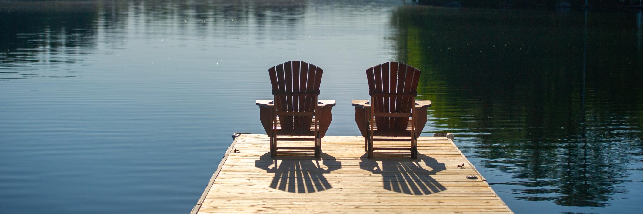 Two Muskoka chairs sitting on a pier over a lake in summertime