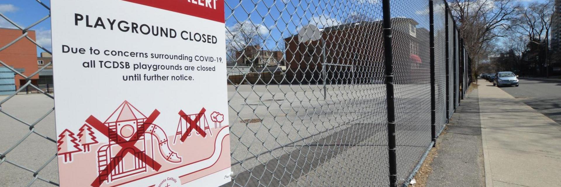 Sign reads Playground Closed, on a chain link fence around a basketball court 