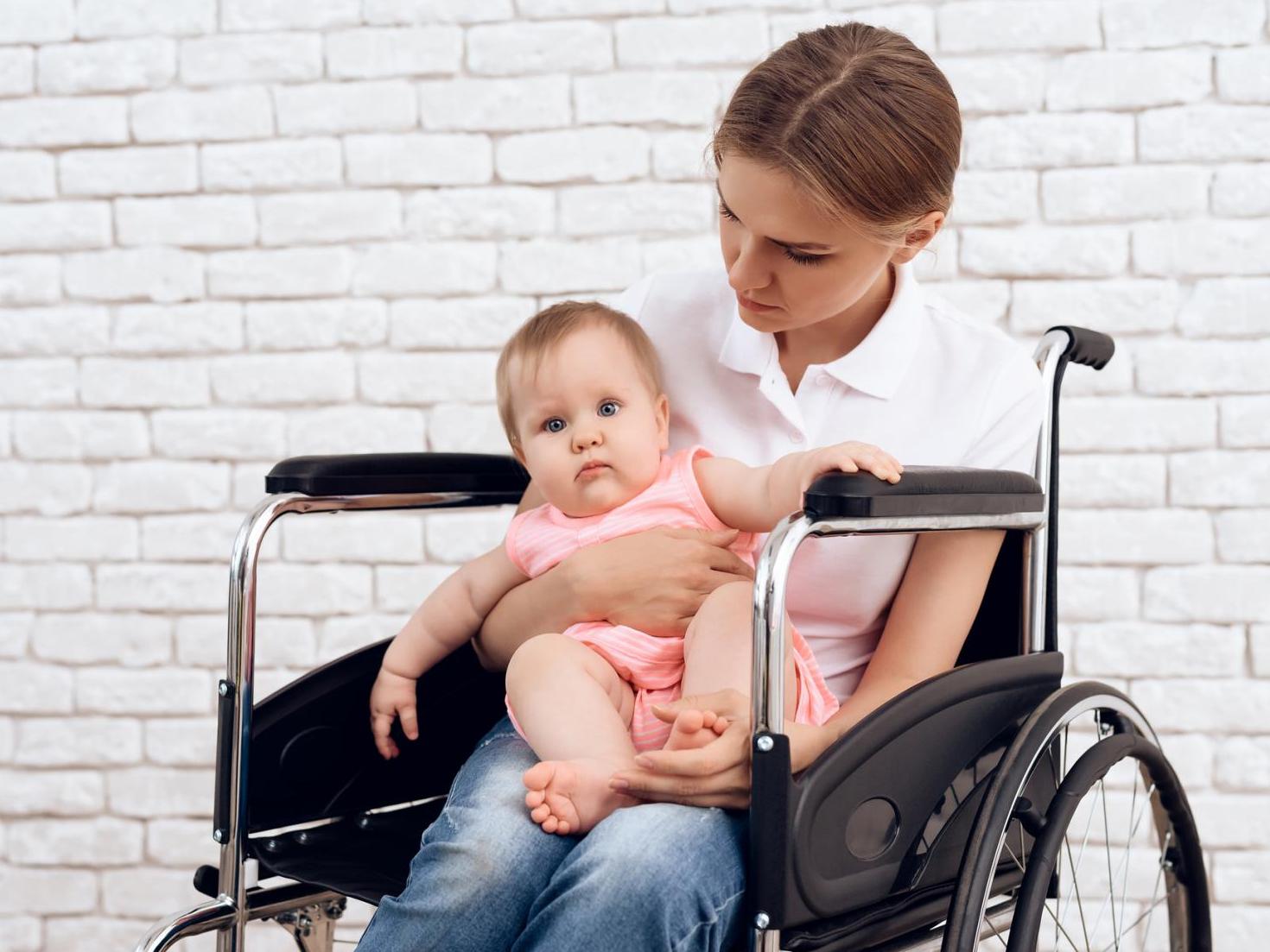 A white woman with blonde hair sits in a wheelchair holding a chubby, healthy baby