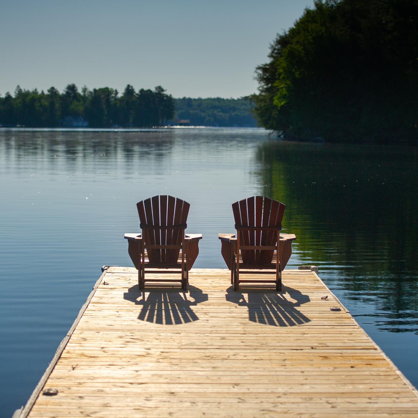Two Muskoka chairs sitting on a pier over a lake in summertime