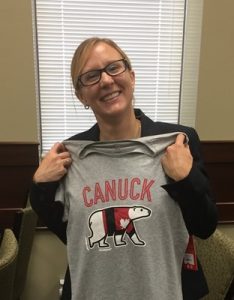A picture of Emma Chan smiling while holding a grey t-shirt with the word CANUCK on it and a polar bear in a coat. 
