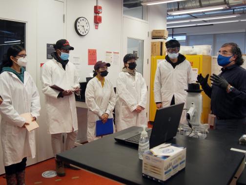 students in lab coats listening to a staff members instructions