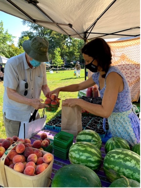 Photo of a vendor at a farmers market handing a customer a small basket of peaches