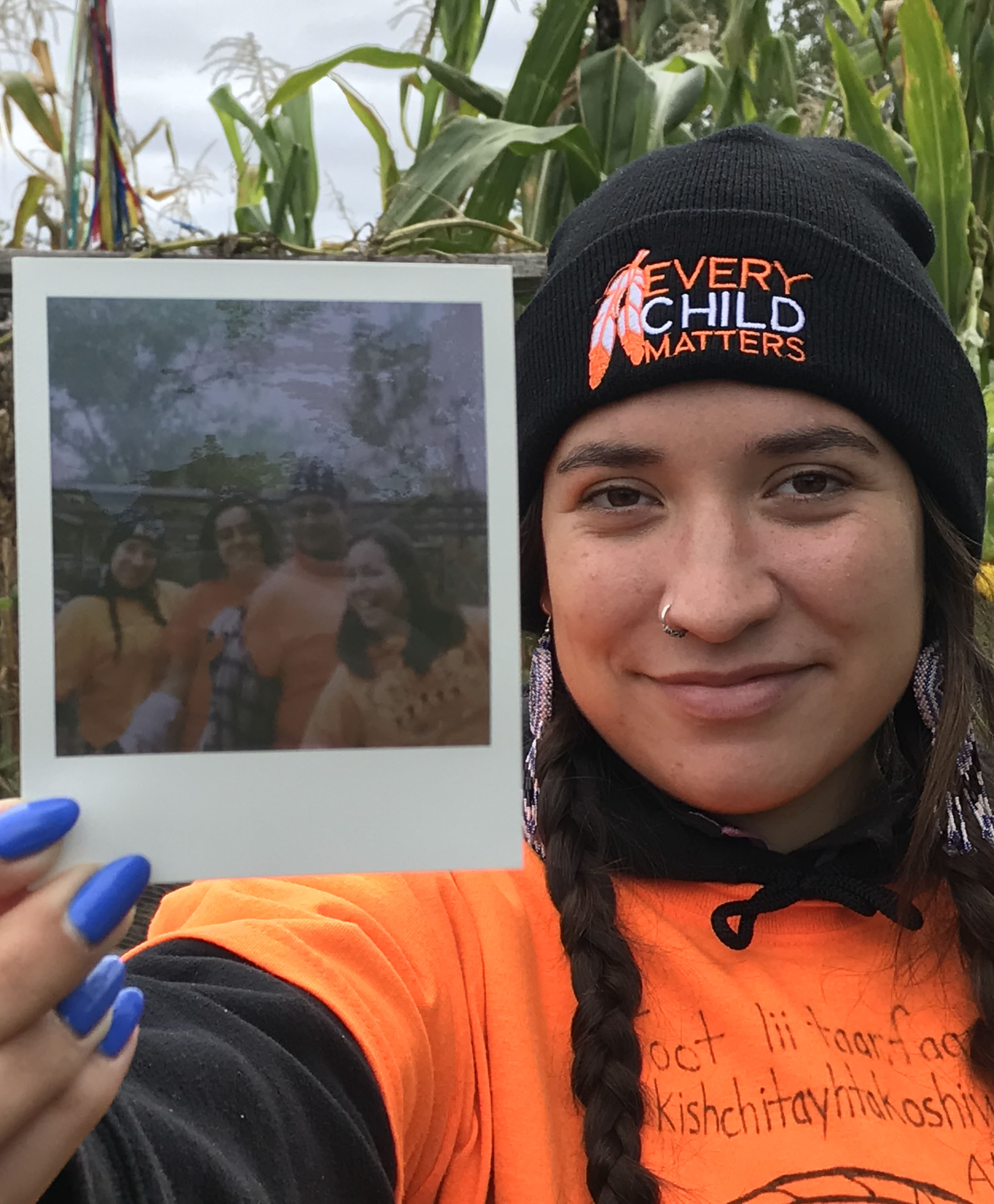 Alexis Bornyk, a young woman with long braided dark hair wearing an "every child matters" hat and orange shirt, holding a polaroid picture from an event at UTSC campus farm