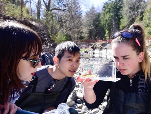 Three students study a fish caught from a river, seen in the background