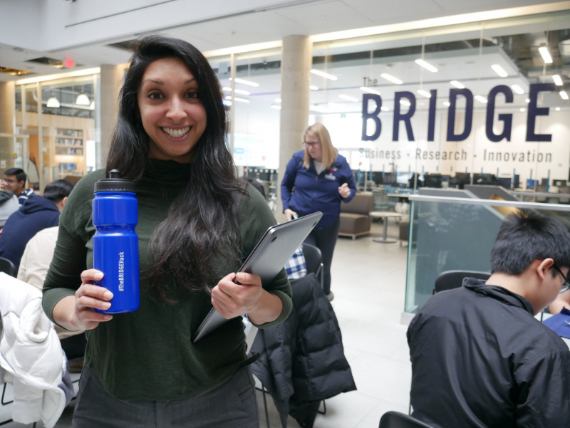 Woman smiling standing near The BRIDGE sign and holding Hackathon bottle