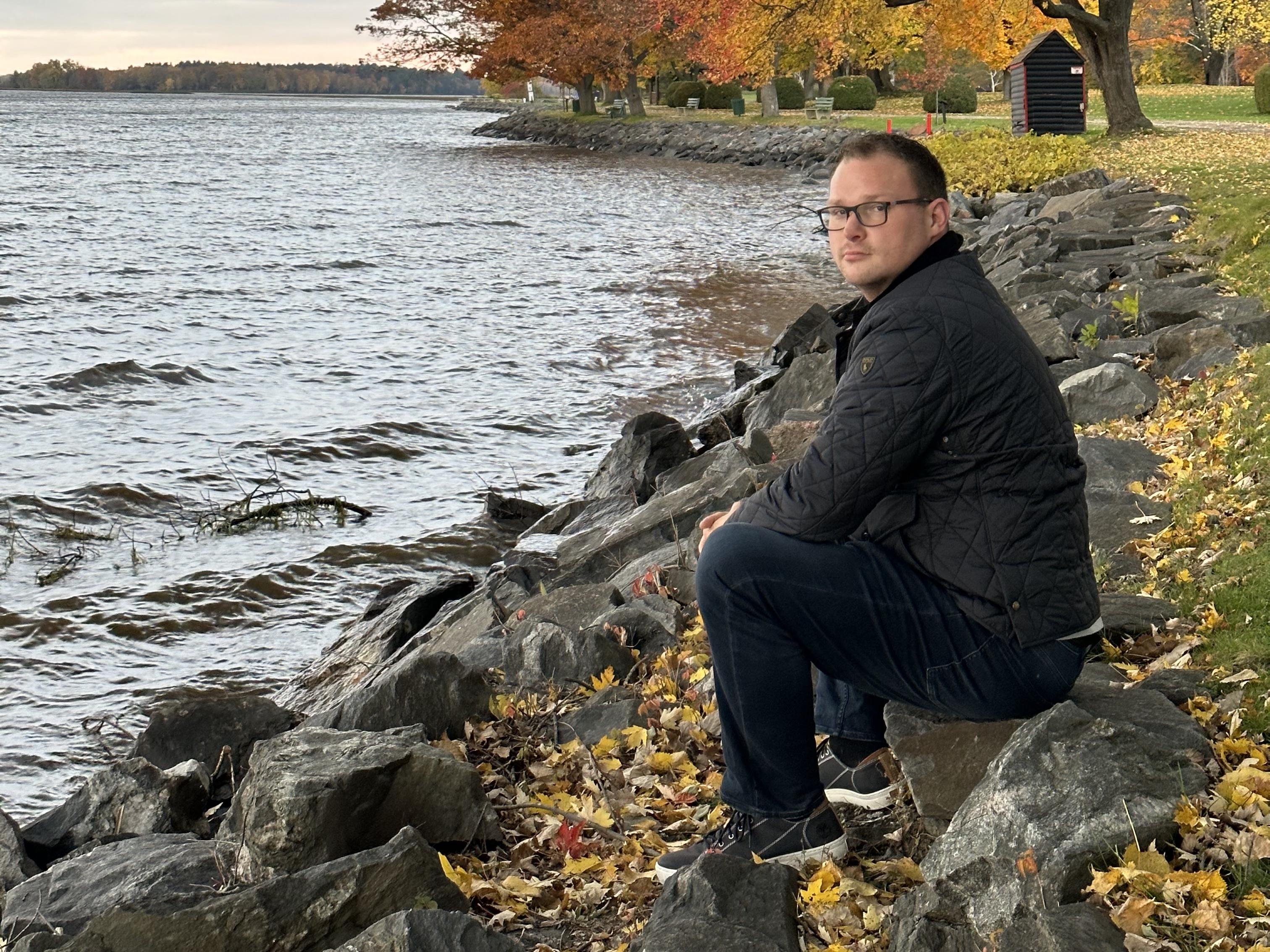 Professor Chad Cowie crouching by a rocky lake shoreline in autumn