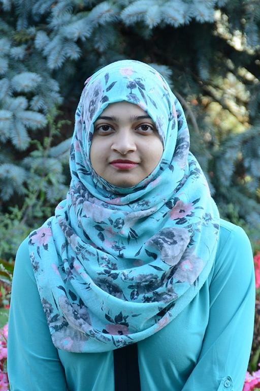 Manaal in a turquoise sweater and hijab standing in a garden in front of a silver spruce